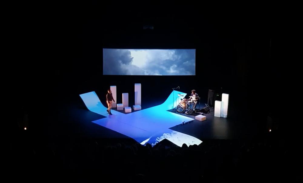 Deux danseurs et un percussionniste sur un plateau blanc, scénographie de blocs clairs.